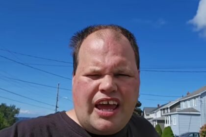 Man in a black shirt speaks to the camera outdoors on a sunny day, with houses and a blue sky in the background.