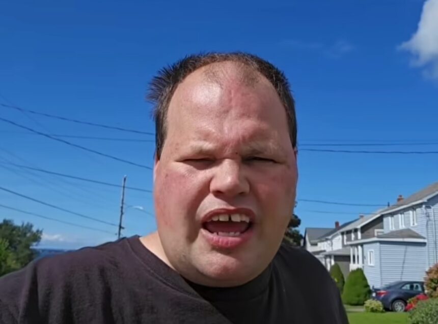 Man in a black shirt speaks to the camera outdoors on a sunny day, with houses and a blue sky in the background.