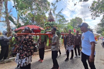 Group of people in floral and patterned clothing carry a tall, decorated centerpiece made of greens and vegetables on a red frame during a street procession.