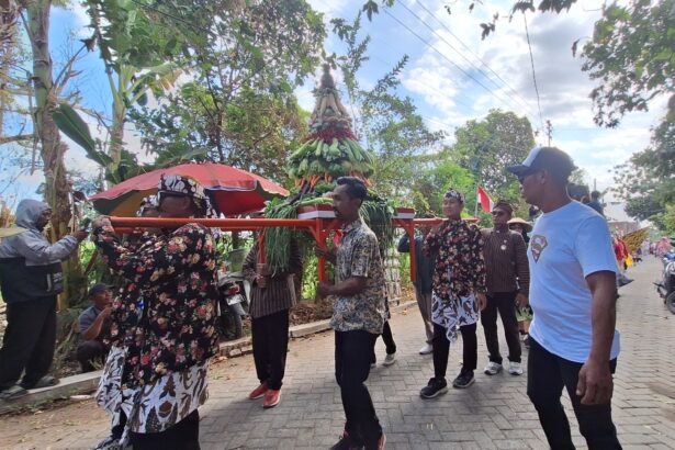 Group of people in floral and patterned clothing carry a tall, decorated centerpiece made of greens and vegetables on a red frame during a street procession.