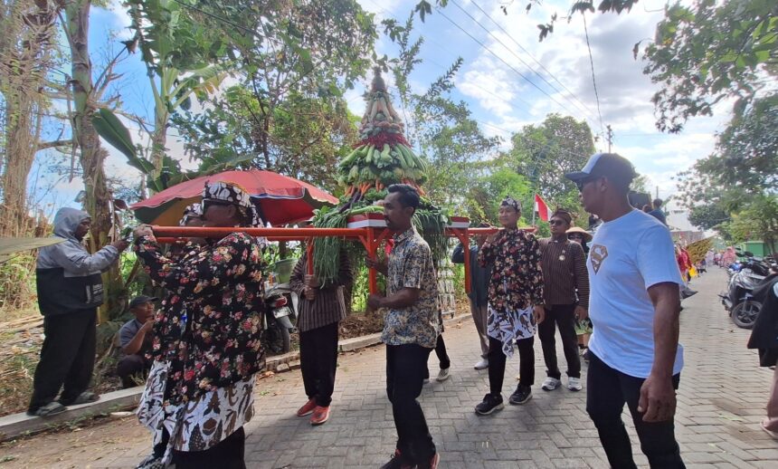 Group of people in floral and patterned clothing carry a tall, decorated centerpiece made of greens and vegetables on a red frame during a street procession.