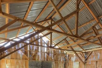 Rustic rural classroom with wooden desks and benches, chalkboard on the wall, and sunlight filtering through gaps in the wooden walls.