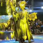 Costumed performer in a large yellow dragon-like suit with horns, holding a staff and a banner on a stage with cheering crowd.