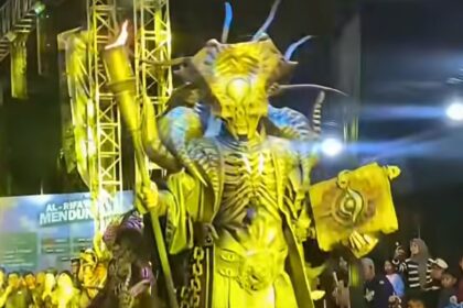 Costumed performer in a large yellow dragon-like suit with horns, holding a staff and a banner on a stage with cheering crowd.