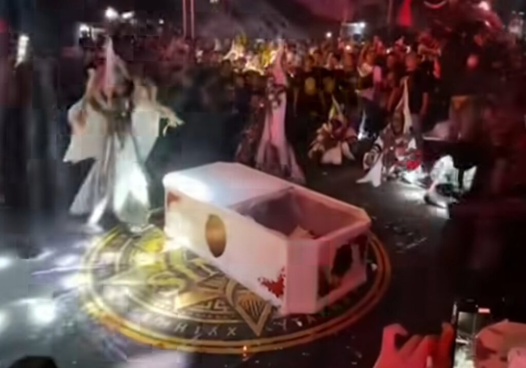 A white coffin-like prop on a glowing circular emblem on the floor, surrounded by performers in white robes amid a night crowd.