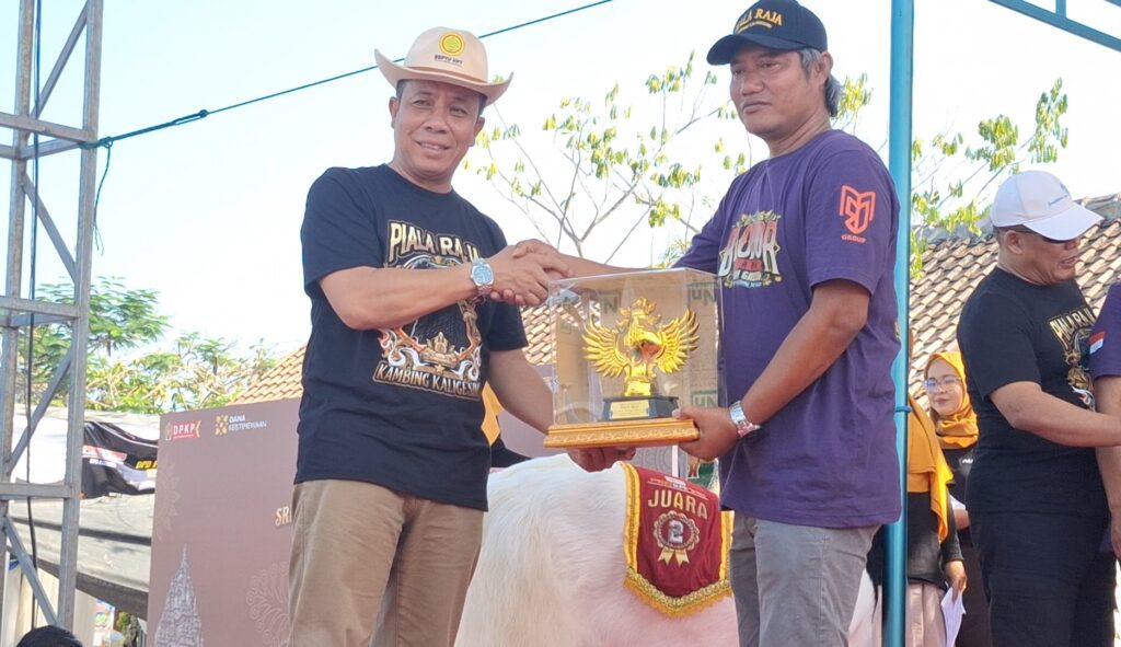 Two men shake hands on stage as one hands the other a clear trophy case with a gold eagle trophy inside. A crowd watches in the background.