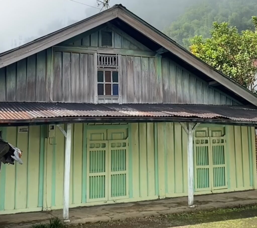 A pale green wooden colonial-style house with a slanted metal roof and a small upper window, set among trees and misty hills in the background.