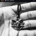 Metal ram-skull pendant on a black cord resting in a hand, shown in black and white.