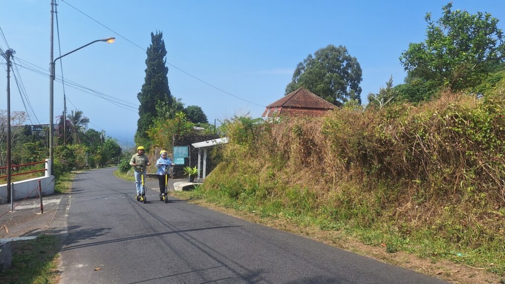 Screenshot_20260917_010608_Gallery - Mabur.co Two people wearing helmets ride yellow electric scooters along a quiet rural road with tall hedges and blue sky above.
