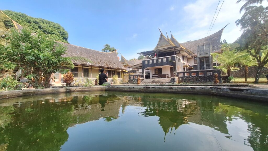 Screenshot_20260917_022527_Gallery - Mabur.co A tranquil temple complex with traditional thatched-roof buildings surrounding a calm, green pond; a person sits by the water's edge.