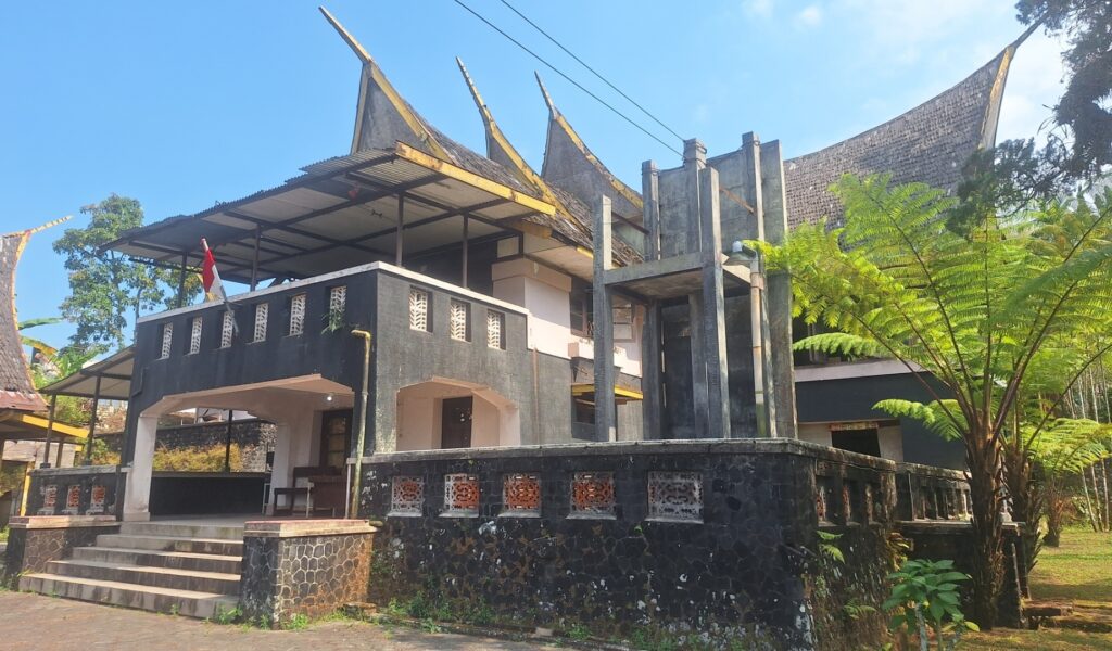 Screenshot_20260917_022600_Gallery - Mabur.co Two-story concrete building with dark exterior, open terrace, and a red-white flag beneath a bright blue sky, surrounded by tropical plants.