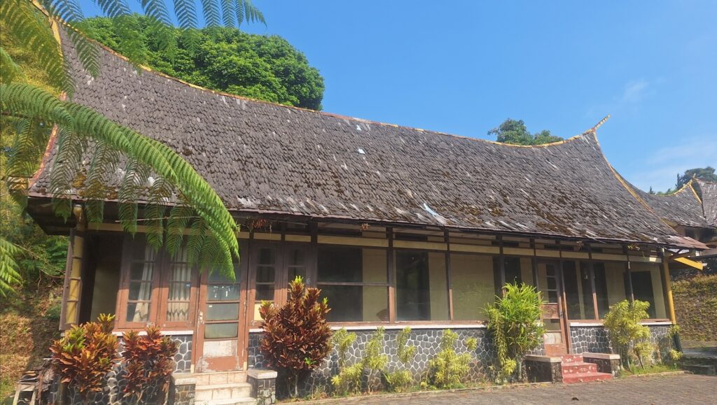 Screenshot_20260917_022622_Gallery - Mabur.co Traditional wooden building with a curved thatched roof, stone base, and tropical plants in front under a blue sky.