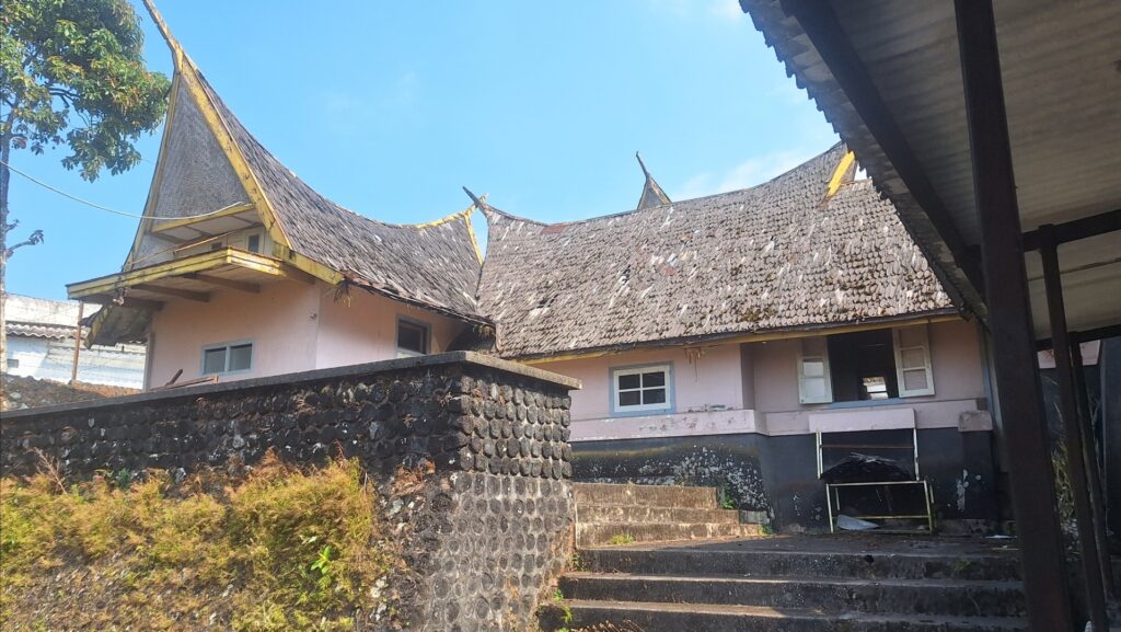 Screenshot_20260917_022646_Gallery - Mabur.co Traditional house with a sweeping thatched roof, pink walls, and a stone retaining wall, set against a clear blue sky.