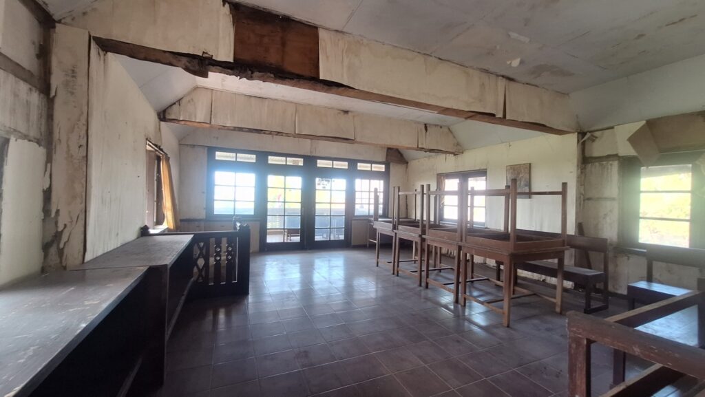 Screenshot_20260917_022845_Gallery - Mabur.co Abandoned room with peeling walls, exposed beams, and stacked wooden tables and benches; large windows at the far end.
