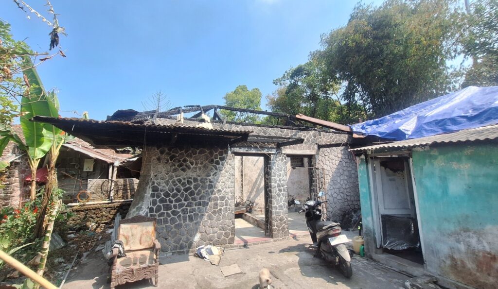 Front view of a stone house with a burnt roof and charred beams, blue tarp draped over part of the structure, and a motorcycle parked nearby in the yard.