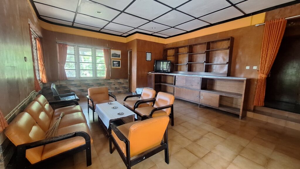 A vintage living room with orange-upholstered chairs around a small table; wood panel walls, tiled floor, and large windows with orange curtains.
