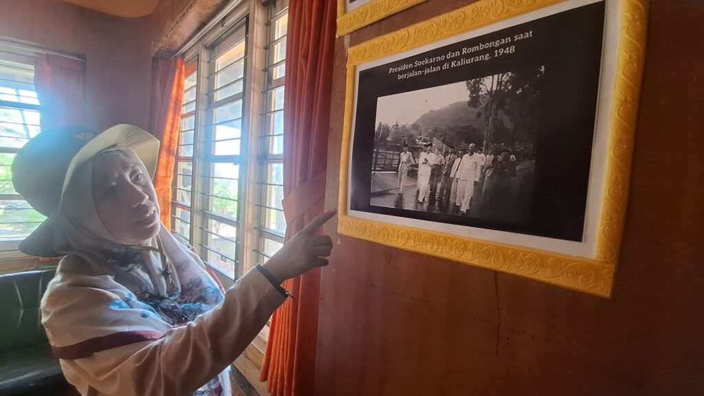 Woman wearing a hat and scarf points at a framed black-and-white historical photo on a wooden wall.