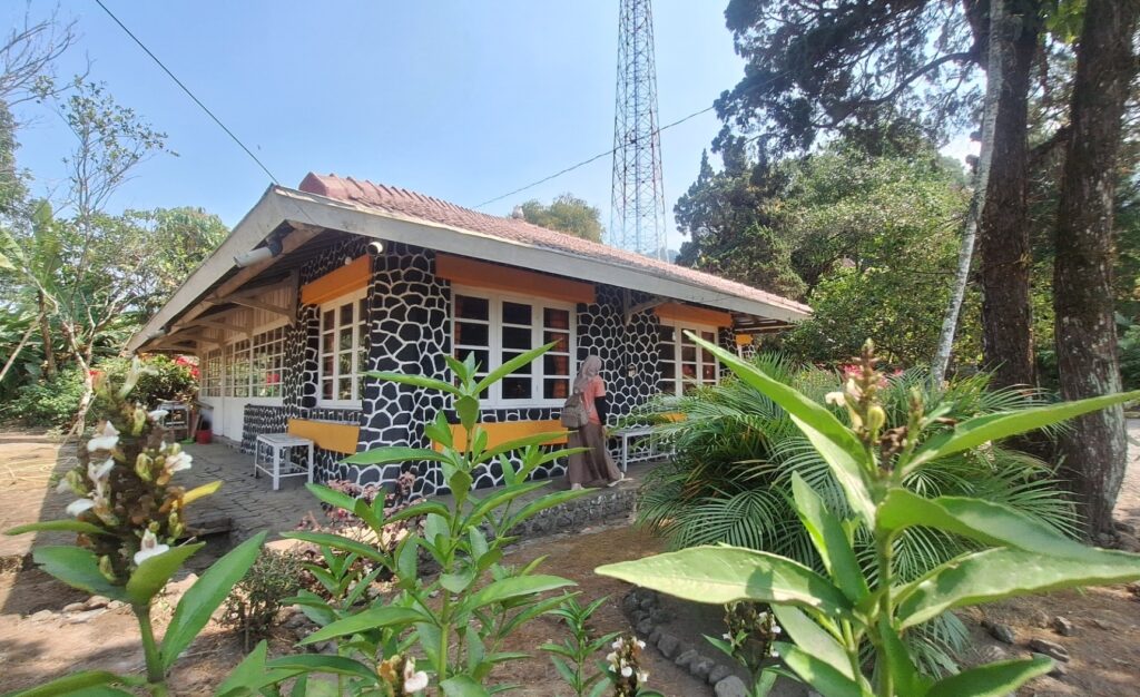 Stone-patterned cottage with orange trim in a tropical garden; a person with a backpack enters the door, sunlight on a sunny day.