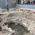 Man performing a ritual by a rocky, circular pit of water; offerings line the rim as onlookers stand behind a gray barrier.