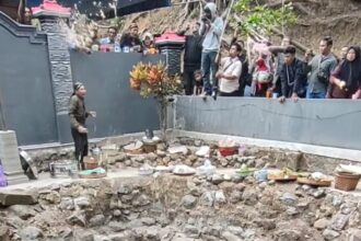 Man performing a ritual by a rocky, circular pit of water; offerings line the rim as onlookers stand behind a gray barrier.
