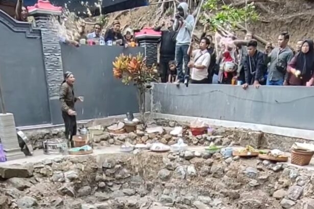 Man performing a ritual by a rocky, circular pit of water; offerings line the rim as onlookers stand behind a gray barrier.