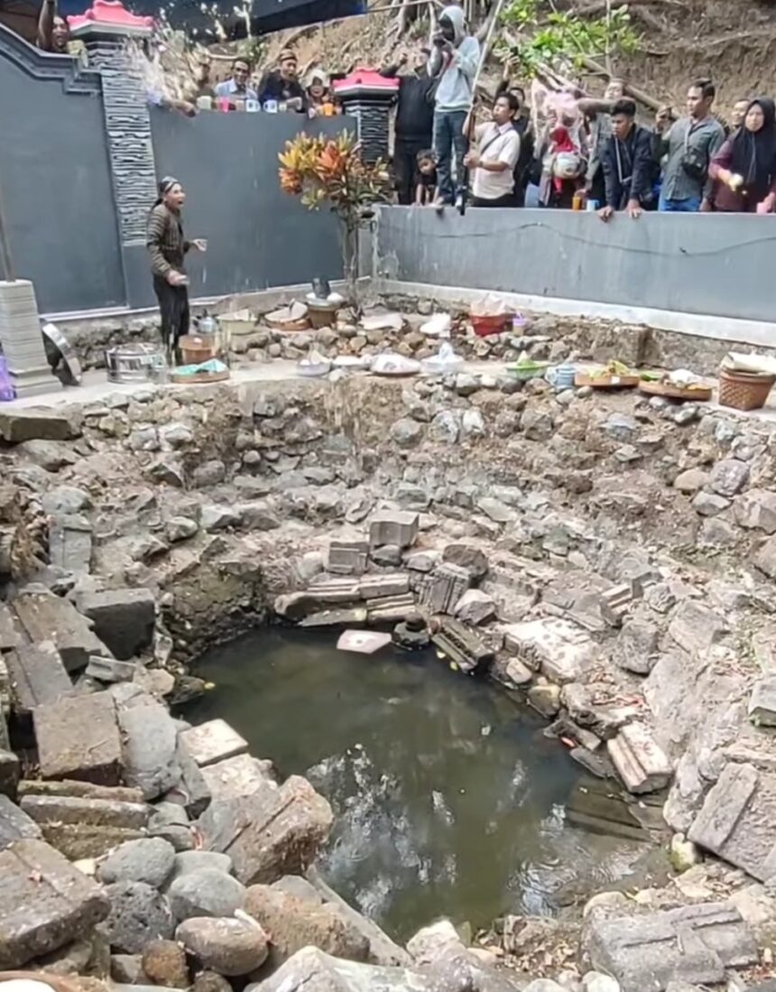 Man performing a ritual by a rocky, circular pit of water; offerings line the rim as onlookers stand behind a gray barrier.