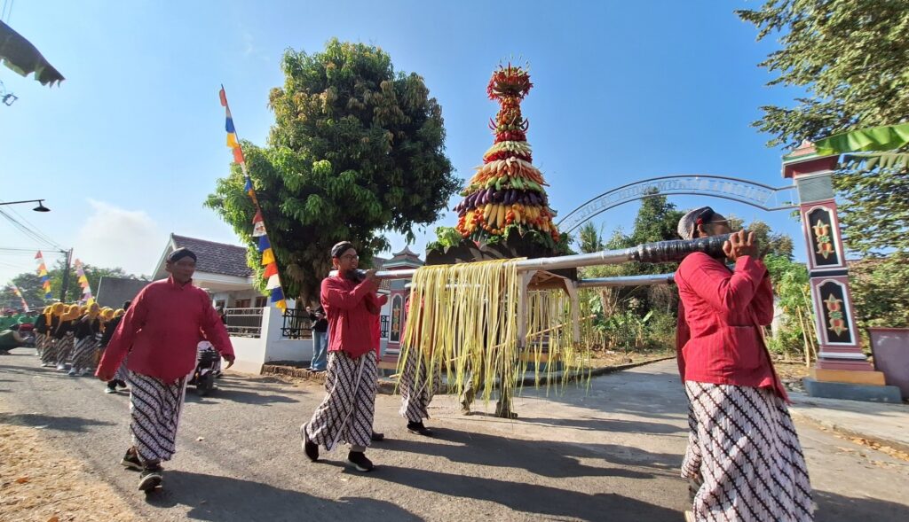 Festival parade with people in red shirts and patterned skirts carrying a tall fruit-and-vegetable sculpture on a pole.