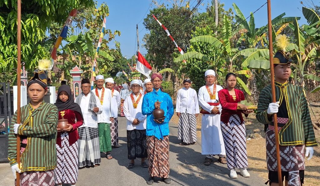 Group of people in traditional clothing marching in a sunny street, carrying ceremonial containers and flags, with lush trees in the background.