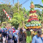 Colorful fruit-and-vegetable sculpture carried in a street festival, with onlookers in traditional clothing nearby.