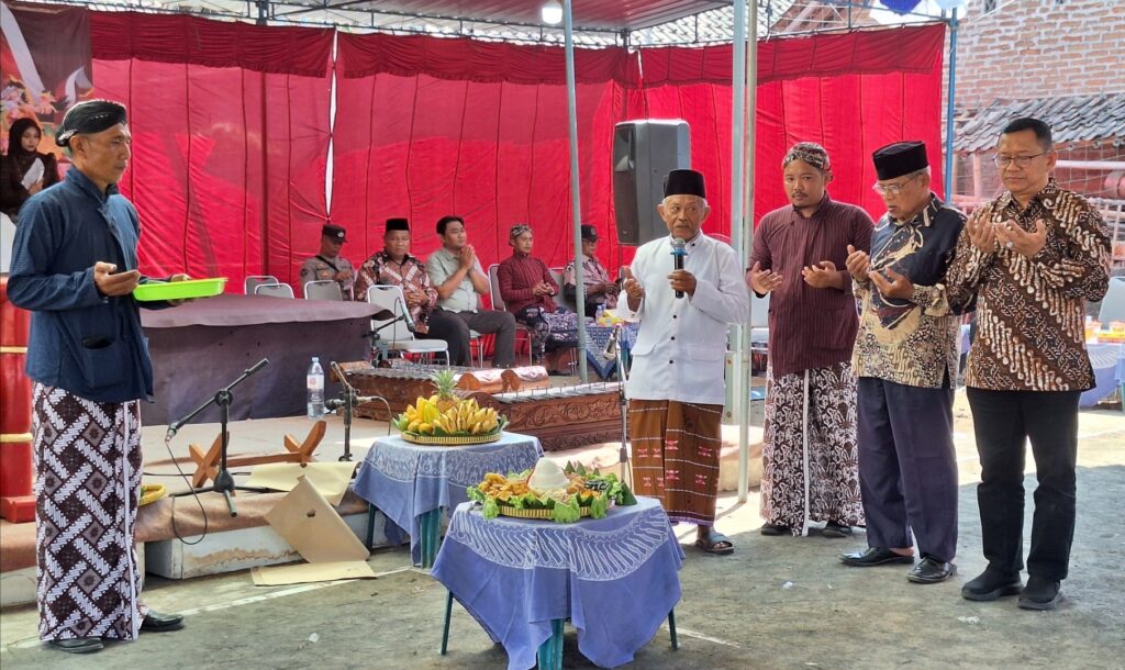 Man with a microphone leading a prayer as others stand with hands raised under a red canopy, traditional outfits visible.