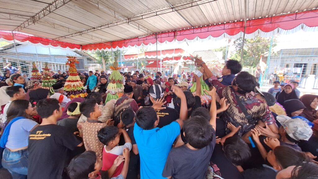 Crowd at an outdoor festival under a red-and-white canopy surrounding tall stacked fruit sculptures on display.