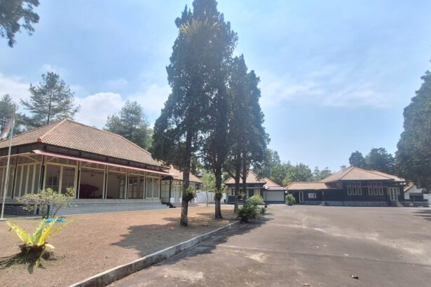 Open courtyard with single-story buildings and red-tiled roofs surrounded by tall trees under a blue sky; a flagpole stands near the left building.