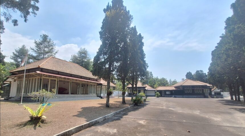Open courtyard with single-story buildings and red-tiled roofs surrounded by tall trees under a blue sky; a flagpole stands near the left building.