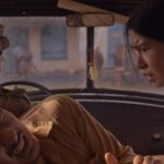 A man in the passenger seat grimaces in pain as a woman in a floral shirt leans in from the right, while the driver sits with his back to the camera in a car interior.