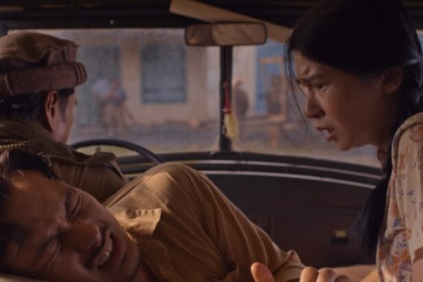 A man in the passenger seat grimaces in pain as a woman in a floral shirt leans in from the right, while the driver sits with his back to the camera in a car interior.