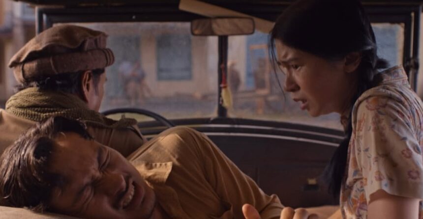 A man in the passenger seat grimaces in pain as a woman in a floral shirt leans in from the right, while the driver sits with his back to the camera in a car interior.