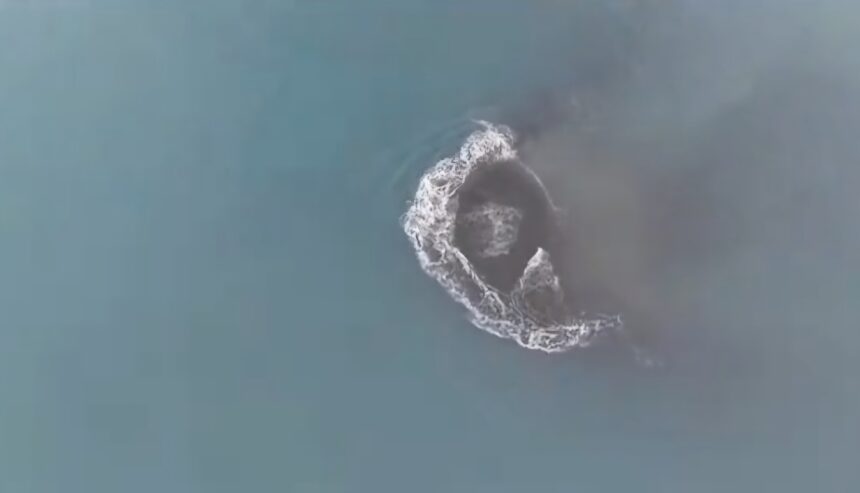 Aerial view of a circular whirlpool or foam ring forming on blue-green ocean water, seen from above.