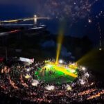 Nighttime aerial view of a circular stadium filled with spectators; performers on a brightly lit green and yellow field with fireworks overhead.