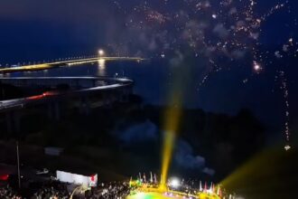 Nighttime aerial view of a circular stadium filled with spectators; performers on a brightly lit green and yellow field with fireworks overhead.