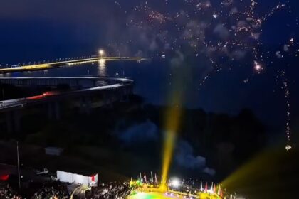 Nighttime aerial view of a circular stadium filled with spectators; performers on a brightly lit green and yellow field with fireworks overhead.