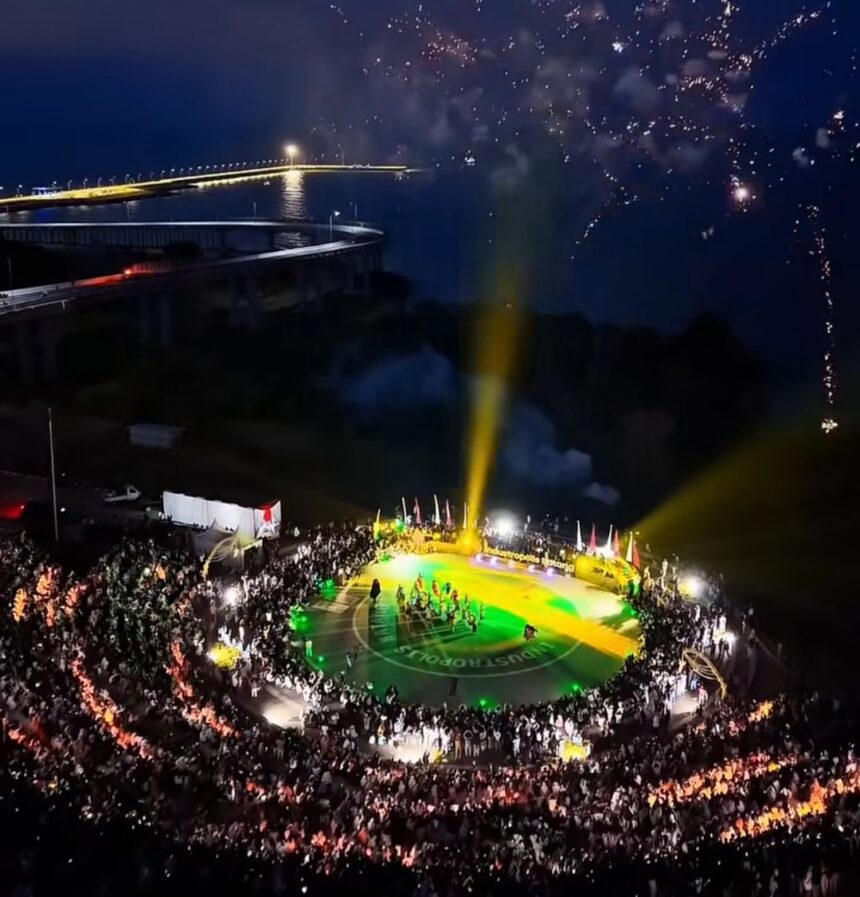 Nighttime aerial view of a circular stadium filled with spectators; performers on a brightly lit green and yellow field with fireworks overhead.