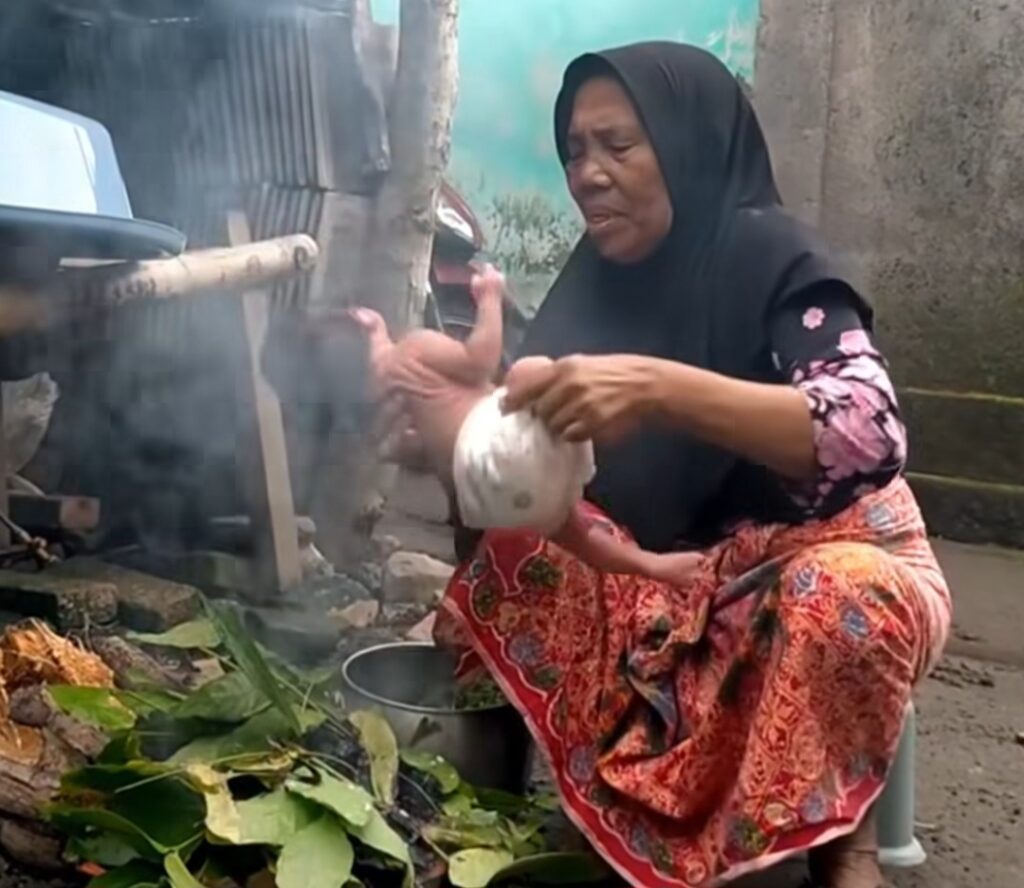 Elderly woman in a black hijab sits by an outdoor wood-fire stove, holding a white pot as steam rises from the pot.