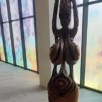 Wooden carved statue in a hallway with colorful frosted windows, figure raising arms overhead with intricate swirling patterns