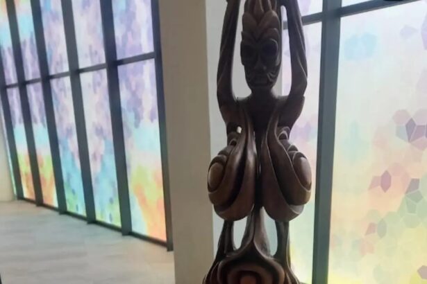 Wooden carved statue in a hallway with colorful frosted windows, figure raising arms overhead with intricate swirling patterns