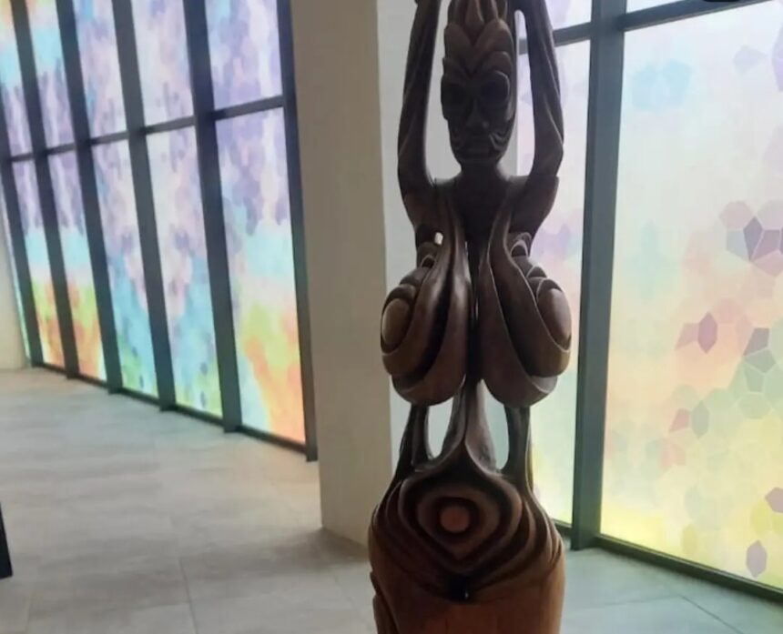 Wooden carved statue in a hallway with colorful frosted windows, figure raising arms overhead with intricate swirling patterns