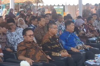 Group of people in batik shirts seated at an outdoor event, listening attentively under a tent.