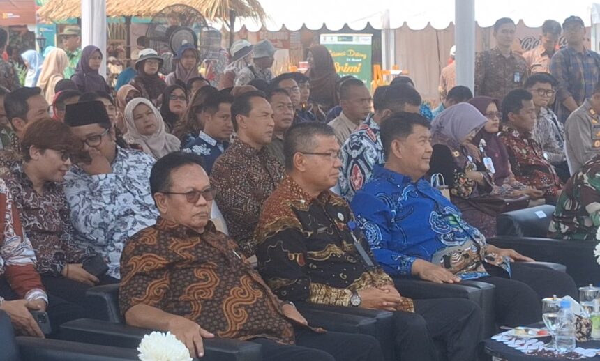 Group of people in batik shirts seated at an outdoor event, listening attentively under a tent.