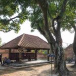 Historic courtyard featuring a small tiled-roof pavilion, brick walls, and large tree roots casting shade.