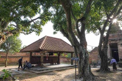 Historic courtyard featuring a small tiled-roof pavilion, brick walls, and large tree roots casting shade.