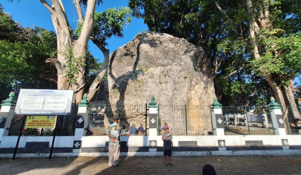 Large rocky boulder behind a decorative green-and-white fence, with trees surrounding it on a sunny day.